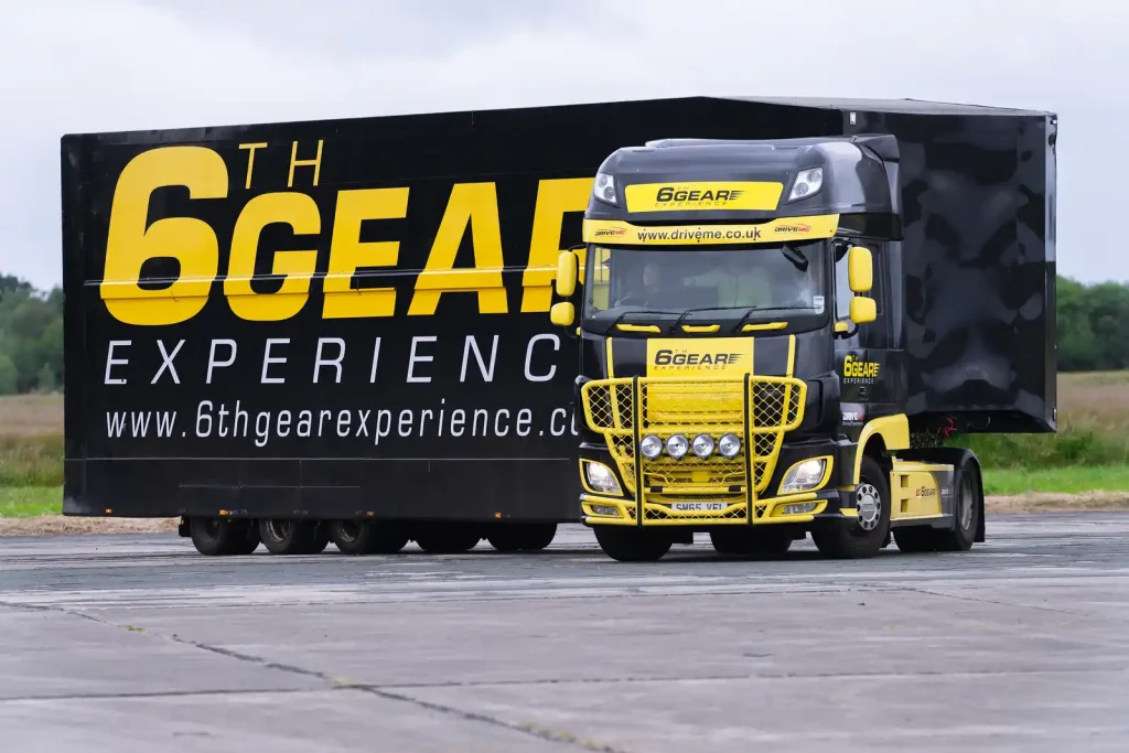 A black and yellow truck with the branding "6th Gear Experience" is parked on a paved surface. The truck has prominent yellow detailing on the front grille and bumper, with additional lights mounted on the front. The side of the truck features large yellow text reading "6th GEAR EXPERIENCE" and the website "www.6thgearexperience.com". The background shows a grassy field and some trees under an overcast sky.